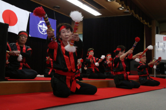 高齢者たちが赤と黒の衣装に身を包み、銭太鼓を披露している写真