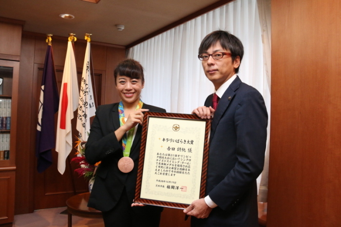 市長室で受賞したキラリいばらき大賞の額入り表彰状を持った茨木市市長と銅メダルを首に掛けた吉田選手の画像